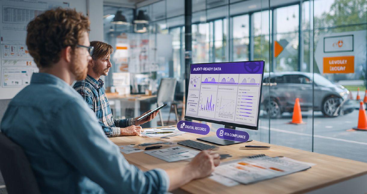 Two professionals in a modern driving centre office analyzing digital audit data on a computer screen labeled “Audit-Ready Data.” The display shows performance charts and compliance metrics, emphasizing digital record management and RTA compliance. Traffi